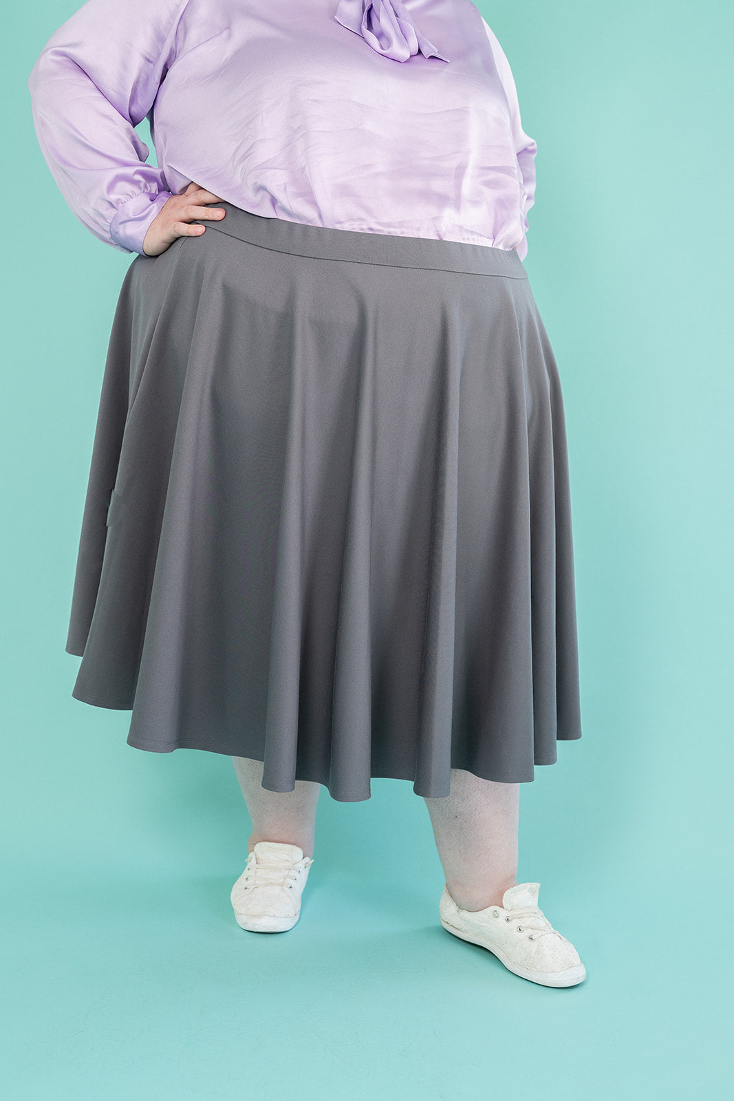 Photo of plus-size woman from shoulders down; she is wearing a lilac coloured blouse and grey skirt with white running shoes
