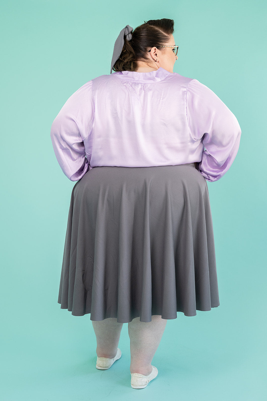 Photo of plus-size brunette woman in lilac coloured blouse and grey skirt. She is facing away from the camera with her hands on her hips.