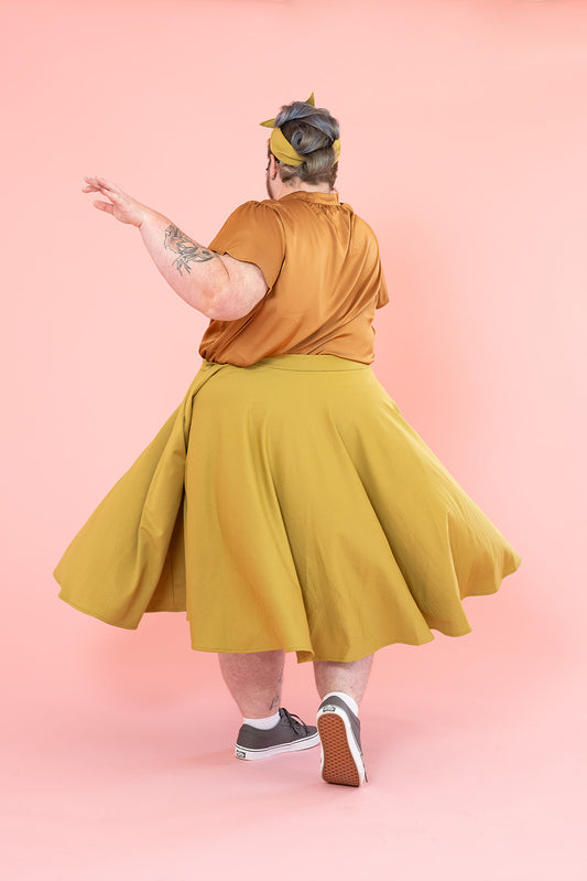 A person is twirling in their mustard-coloured skirt. They are wearing a bronze silk shirt and have several tattoos.