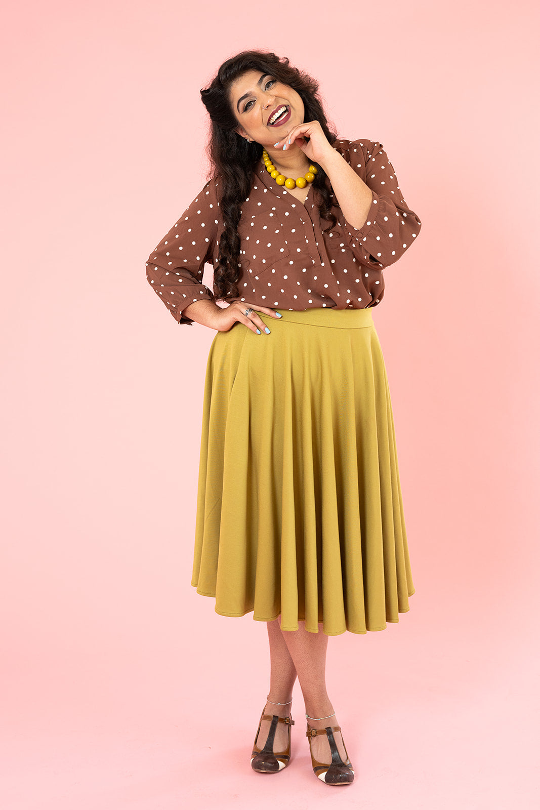 A woman with dark brown hair is looking at the camera and smiling. She is wearing a mustard coloured skirt and a brown shirt with polka dots