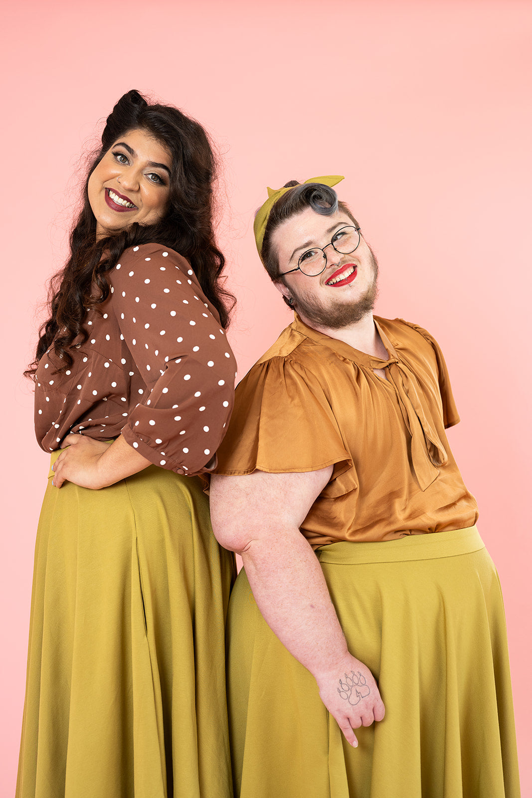 Photo of two people standing back to back and wearing mustard colour shirts. One is a woman with long brown hair and is wearing a brown shirt. The other person is wearing a bronze silk shirt and has facial hair and red lipstick